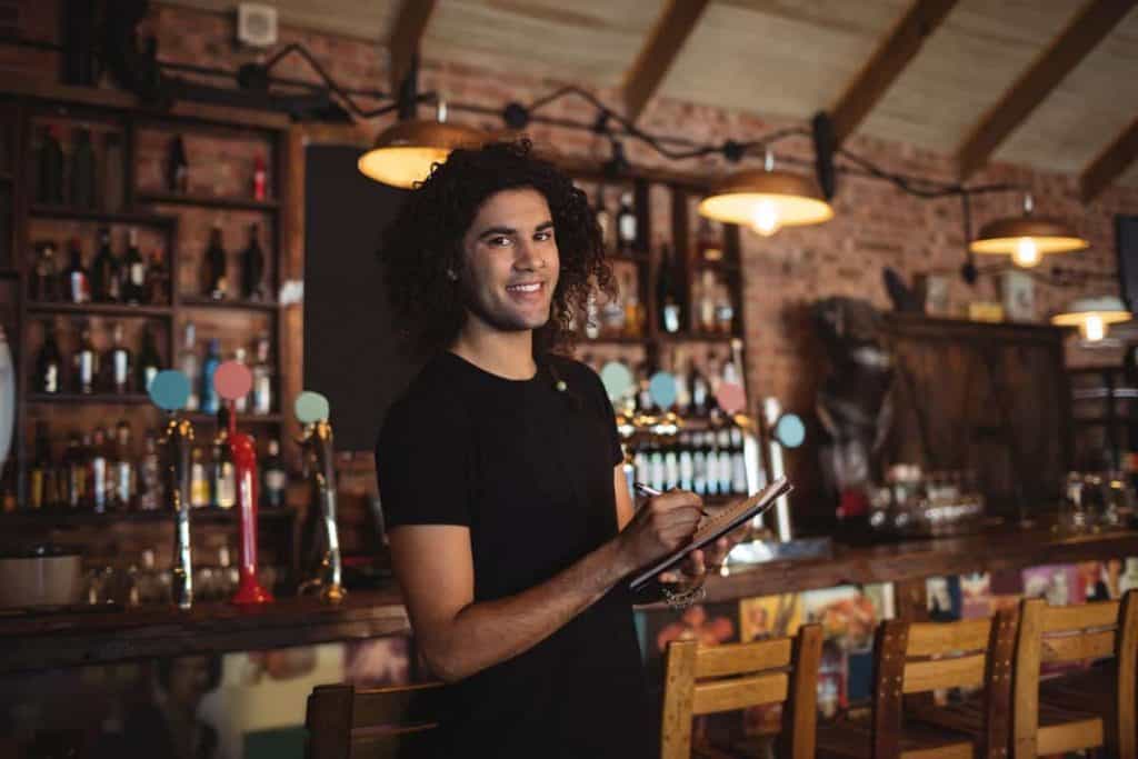 Ein lächelnder Kellner mit lockigem Haar steht mit Notizblock und Stift in der Hand in einer rustikalen Bar, im Hintergrund sind Regale mit Flaschen und warme Hängelampen zu sehen.