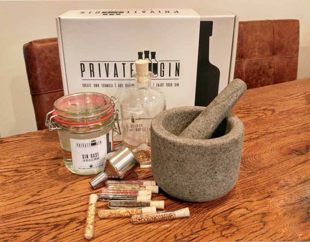 Auf einem Holztisch steht ein Set zur Gin-Herstellung, bestehend aus einer Flasche, einem mit „Gin Base“ beschrifteten Glas, Reagenzgläsern mit Botanicals, einem Metalltrichter sowie einem Mörser und Stößel aus Stein. Im Hintergrund stehen braune Ledersessel.