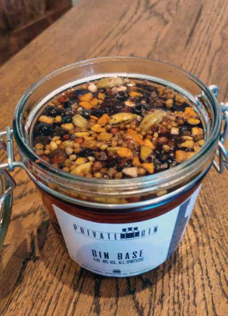 Ein Glasgefäß mit der Aufschrift „Private Gin Gin Base“ steht auf einem Holztisch; es ist mit einer dunklen Flüssigkeit gefüllt, in der Gewürzstücke und Pflanzenbestandteile schwimmen.