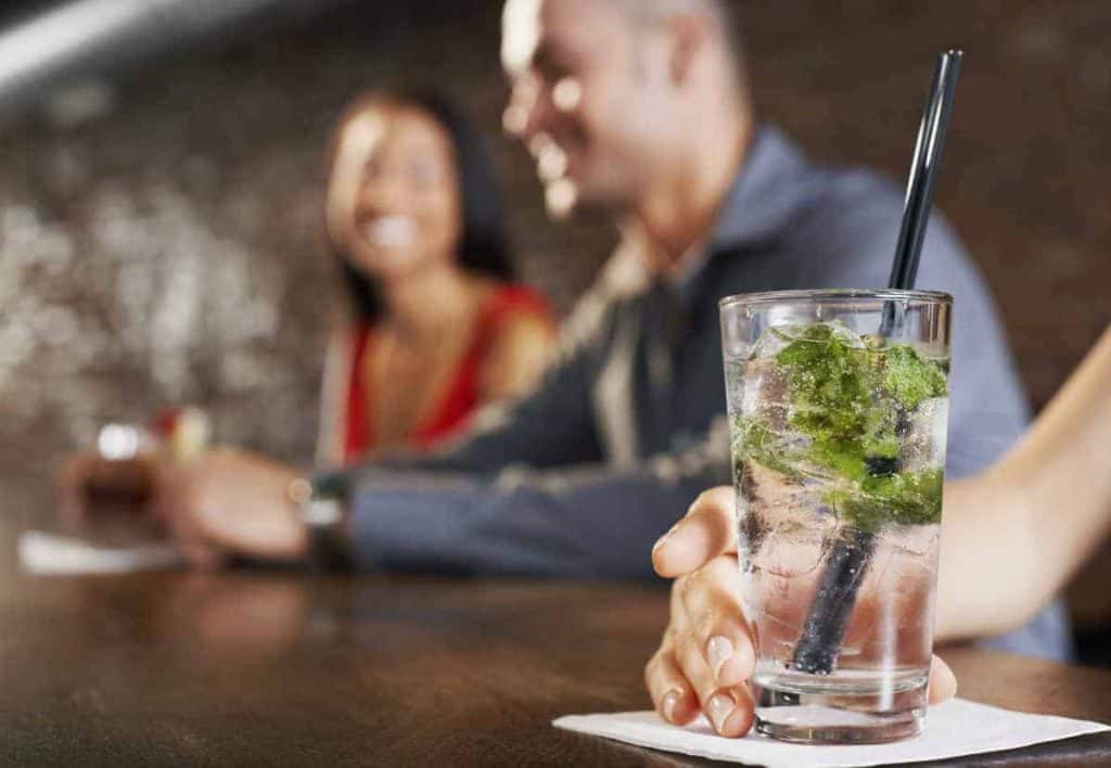 Eine Nahaufnahme eines Mojito-Cocktails mit Minze und einem Strohhalm auf einer Serviette; im unscharfen Hintergrund sind zwei Menschen zu sehen, die in einer Bar lächeln und sich unterhalten.