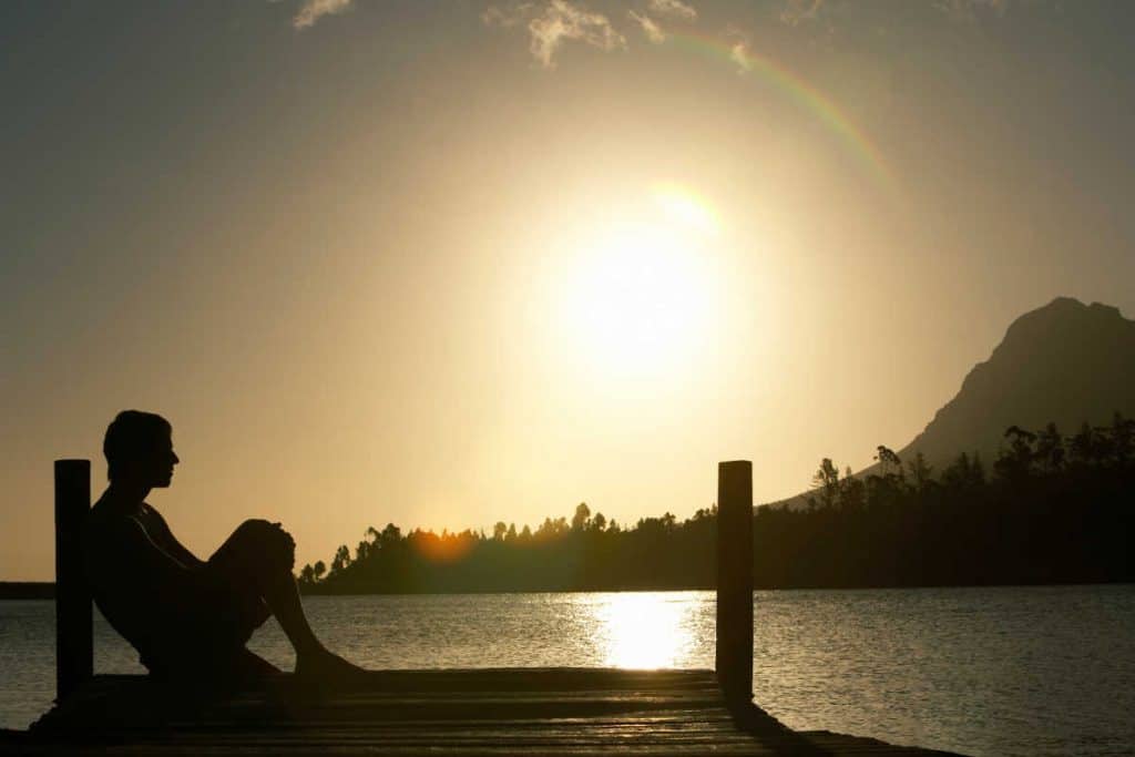 Silhouette einer Person, die bei Sonnenuntergang auf einem hölzernen Steg an einem ruhigen See sitzt, im Hintergrund Berge und Bäume sowie ein schwacher Regenbogen am Himmel.