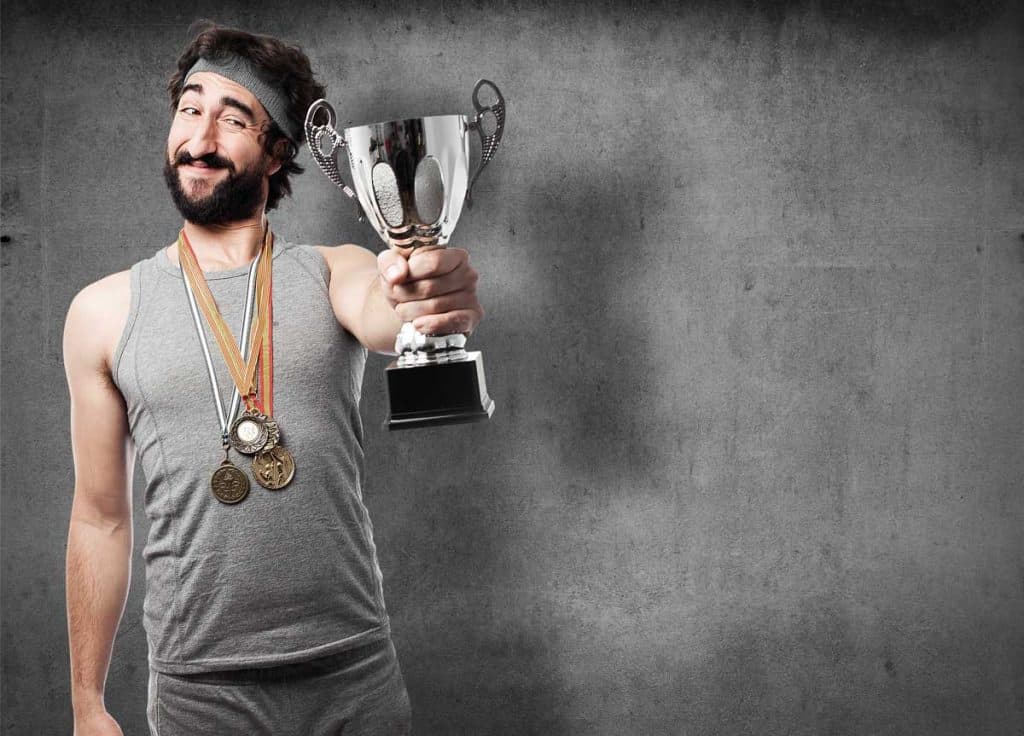 Ein lächelnder Mann in Sportkleidung, der Medaillen um den Hals trägt, hält einen Pokal hoch und posiert stolz vor einem schlichten grauen Hintergrund.