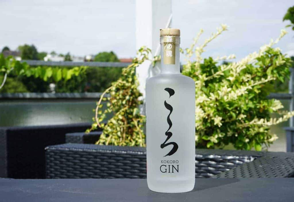 Auf einem schwarzen Gartentisch steht eine mattierte Flasche Kokoro Gin mit japanischen Schriftzeichen, im unscharfen Hintergrund sind Grünpflanzen und Gartenmöbel zu sehen.