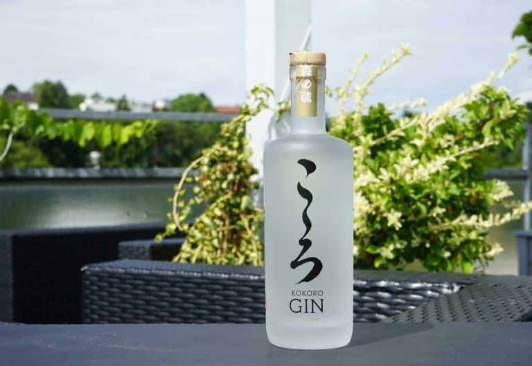 Auf einem schwarzen Gartentisch steht eine mattierte Flasche Kokoro Gin mit japanischen Schriftzeichen, im unscharfen Hintergrund sind Grünpflanzen und Gartenmöbel zu sehen.