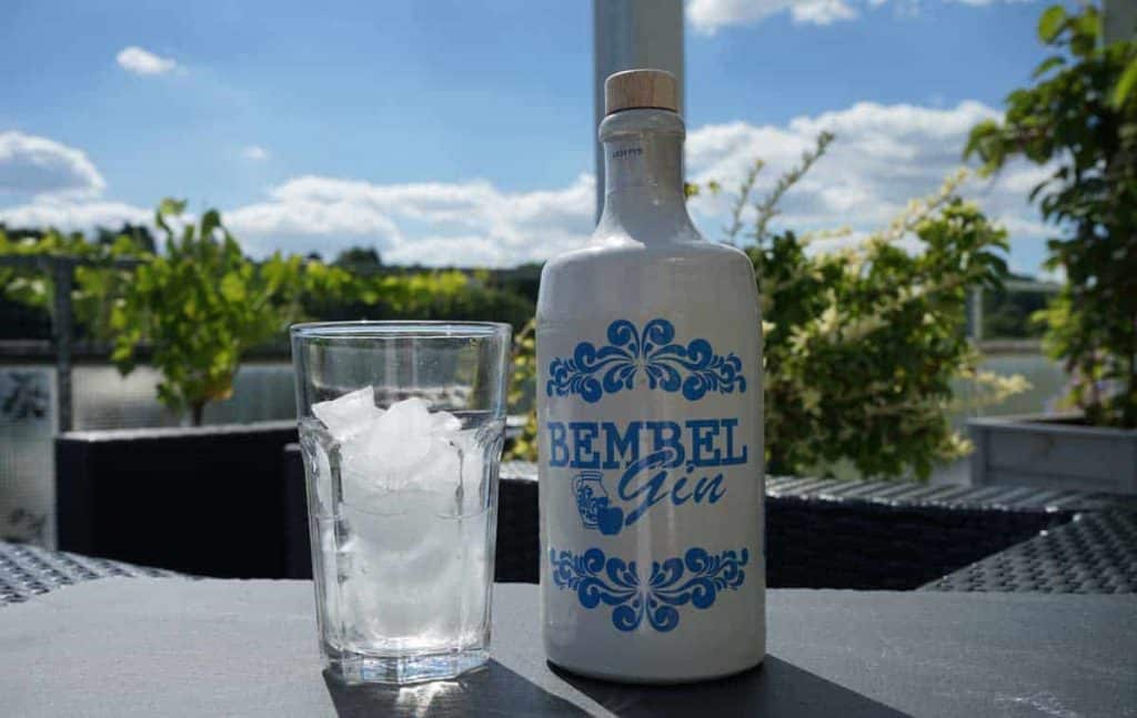 Auf einem Tisch im Freien steht eine Flasche mit der Aufschrift „Bembel Gin“ neben einem mit Eiswürfeln gefüllten Glas, im Hintergrund sind Grünpflanzen und ein blauer Himmel mit Wolken zu sehen.