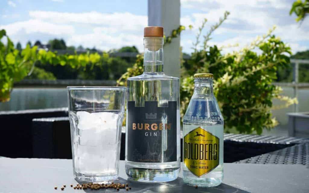 Auf einem Tisch im Freien stehen ein Glas mit Eis, eine Flasche Burgen-Gin und eine Flasche Goldberg-Tonic-Water, im Hintergrund ist ein sonniger Flussuferbereich zu sehen, und in der Nähe stehen grüne Pflanzen.