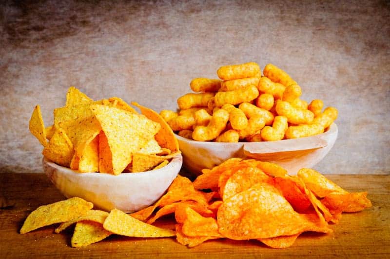 Auf einer Holzplatte stehen zwei Schalen, gefüllt mit gelben Maistortilla-Chips und orangefarbenen Käsebällchen; davor liegt vor einem braunen Hintergrund ein Haufen orangefarbener Kartoffelchips.