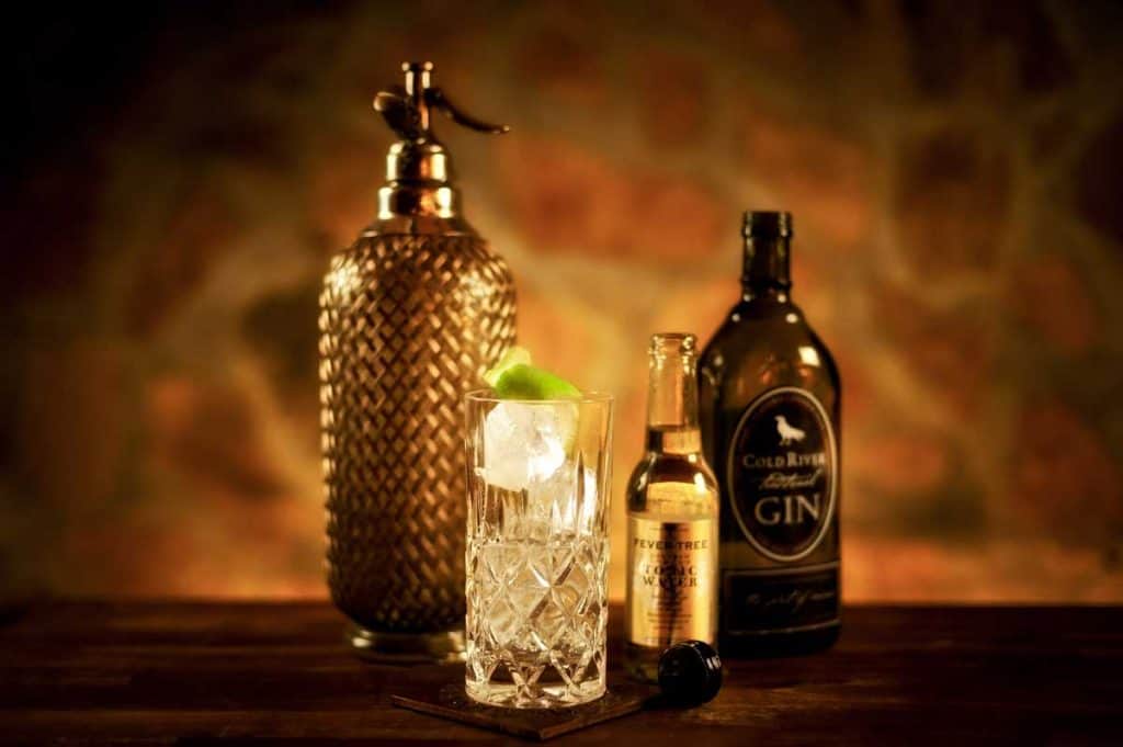 Ein Kristallglas mit Eis und Limette steht neben einer Vintage-Seltzerflasche, einer kleinen Tonic-Flasche und einer dunklen Gin-Flasche auf einer Holzunterlage vor einem warmen, sanft beleuchteten Hintergrund.