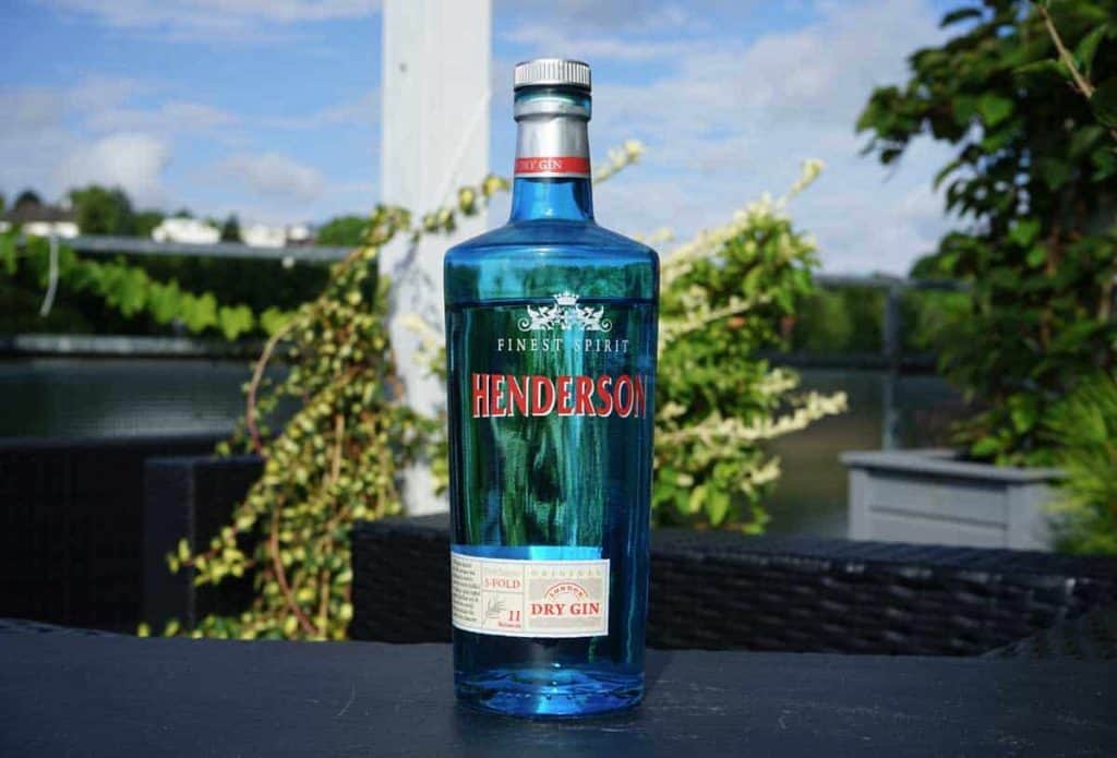 Auf einem Tisch im Freien steht eine blaue Glasflasche mit Henderson Dry Gin, im unscharfen Hintergrund sind Grünpflanzen und ein Zaun zu sehen.