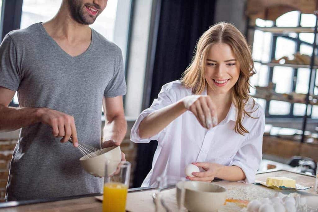 Eine Frau lächelt, während sie ein Ei in eine Schüssel aufschlägt, und ein Mann neben ihr verrührt die Zutaten. Sie kochen gemeinsam in einer hellen Küche, auf der Arbeitsplatte liegen die Zutaten bereit.