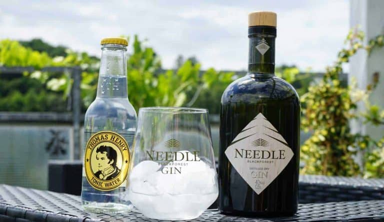 Eine Flasche Thomas Henry Tonic Water, ein Glas mit Eiswürfeln und der Aufschrift „Needle Blackforest Gin“ sowie eine Flasche Needle Blackforest Gin auf einem Korbtisch im Freien, im Hintergrund ist Grün zu sehen.