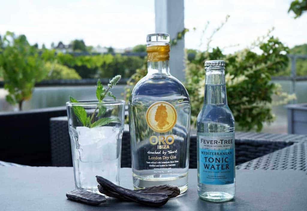 Ein Glas mit Eis und Minze, eine Flasche ORO Ibiza Gin, eine Flasche Fever-Tree Mediterranean Tonic Water und zwei dunkle Kapseln sind auf einem Tisch im Freien vor einem grünen Hintergrund angeordnet.