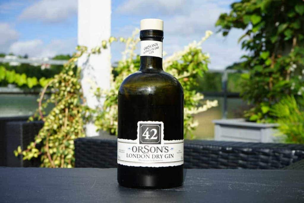 Eine Flasche „Orsons London Dry Gin“ mit einem Etikett, auf dem die Zahl 42 zu sehen ist, steht auf einem Tisch im Freien, im Hintergrund sind Grünpflanzen und ein bewölkter Himmel zu sehen.