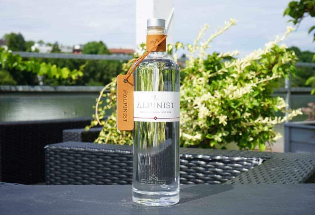 Auf einem schwarzen Tisch im Freien steht eine durchsichtige Flasche Alpinist-Gin mit einem Anhänger am Flaschenhals, im Hintergrund sind Grünflächen und Gebäude verschwommen zu sehen.