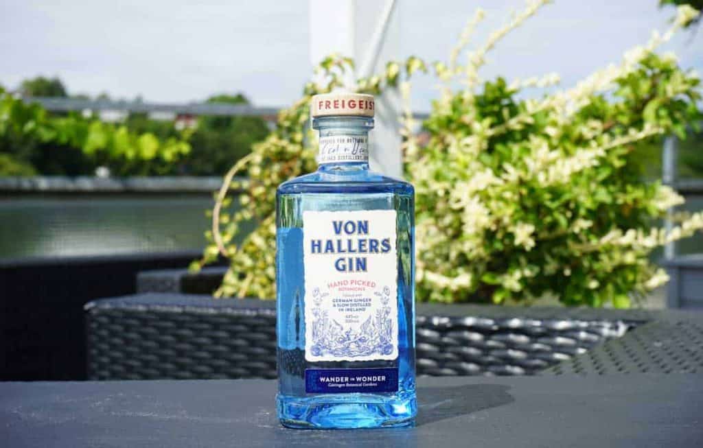 Auf einem Tisch im Freien steht eine durchsichtige Flasche „Von Hallers Gin“, umgeben von Grünpflanzen und vor einem unscharfen Hintergrund aus Pflanzen und einem Metallgeländer.