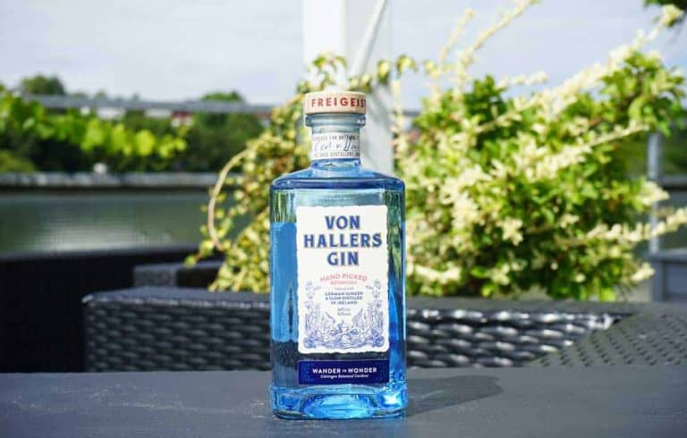 Auf einem Tisch im Freien steht eine durchsichtige Flasche „Von Hallers Gin“, umgeben von Grünpflanzen und vor einem unscharfen Hintergrund aus Pflanzen und einem Metallgeländer.