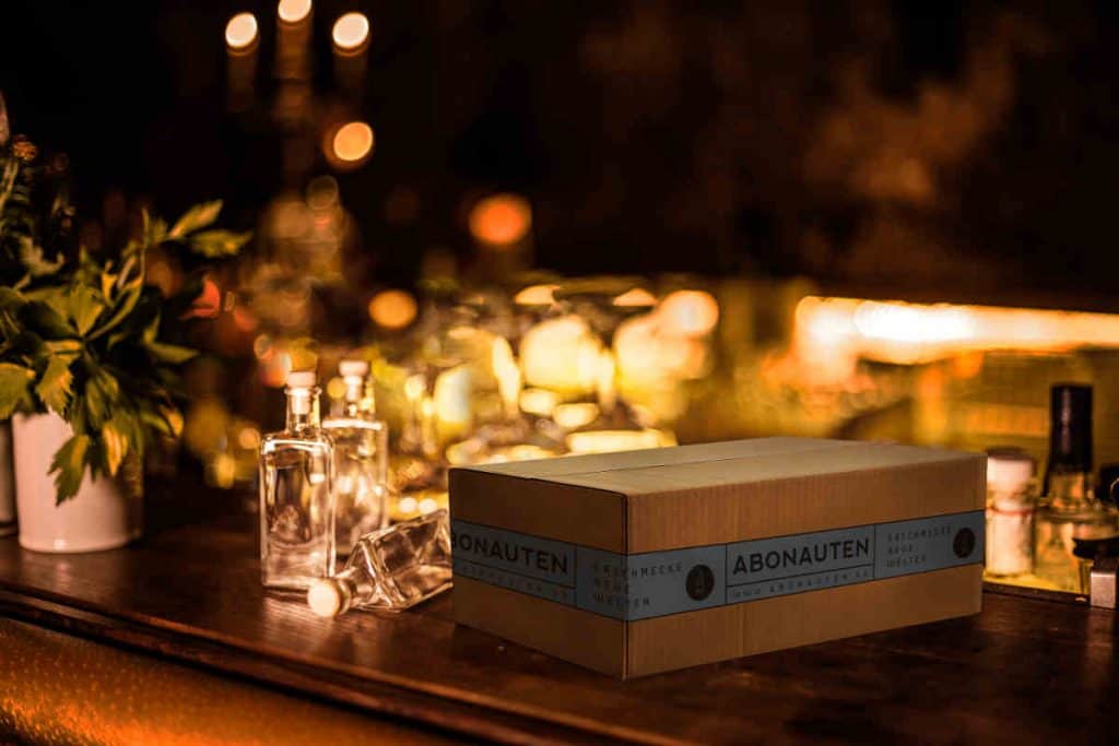 Auf einer Holzleiste steht ein Karton mit der Aufschrift „ABONAUTEN“, umgeben von Glasflaschen und gedämpftem, warmem Licht vor einem verschwommenen, festlichen Hintergrund.