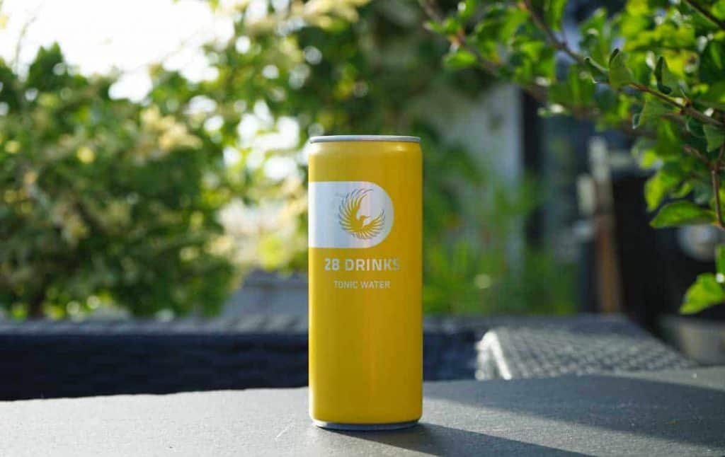Auf einem dunklen Tisch im Freien steht eine gelbe Dose mit der Aufschrift „28 DRINKS TONIC WATER“, umgeben von Grünpflanzen und mit Sonnenlicht im Hintergrund.
