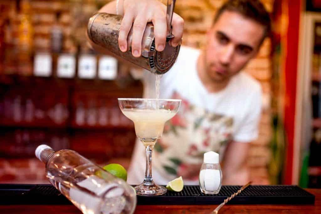 Ein Barkeeper gießt in einer Bar einen Cocktail aus einem Shaker in ein Martini-Glas. Auf der Theke stehen eine Flasche, eine Limette, ein Salzstreuer und eine Limettenscheibe. Der Hintergrund ist unscharf.