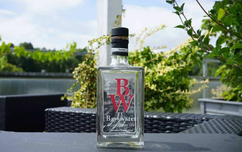 Auf einem Tisch im Freien steht eine Flasche Bayswater London Dry Gin, im Hintergrund sind Grünpflanzen und ein Gewässer zu sehen.