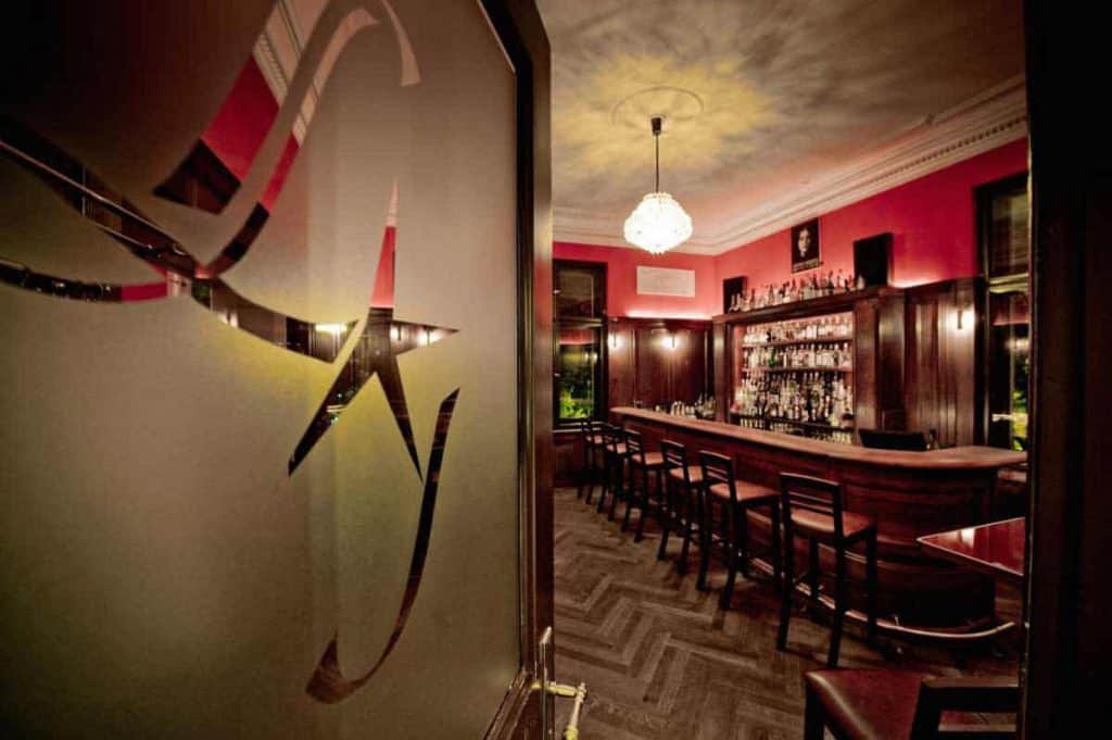 Eine warm beleuchtete, leere Bar mit dunkler Holzausstattung, einer langen Theke mit Barhockern, Flaschenregalen im Hintergrund und einem Kronleuchter, der von der Decke hängt. Im Vordergrund steht eine Milchglastür mit Sternmotiv, die offen ist.