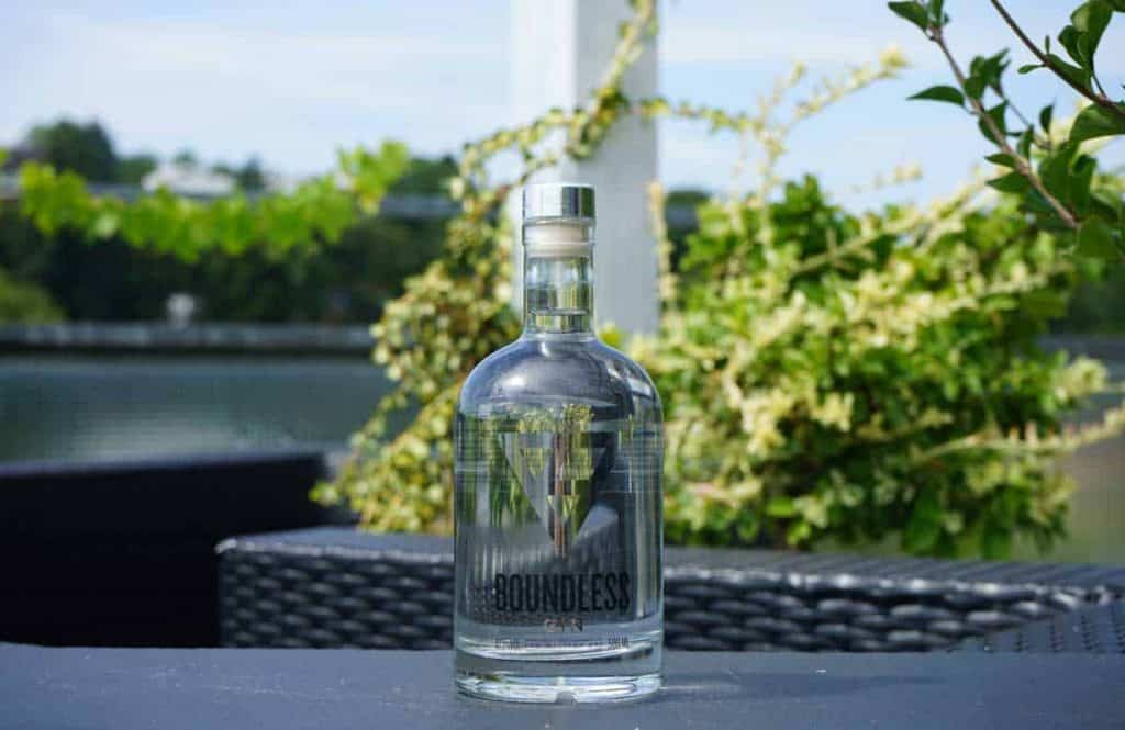 Eine durchsichtige Flasche mit der Aufschrift „Boundless Gin“ steht im Freien auf einer dunklen Fläche, im unscharfen Hintergrund sind grüne Pflanzen und Wasser zu sehen.