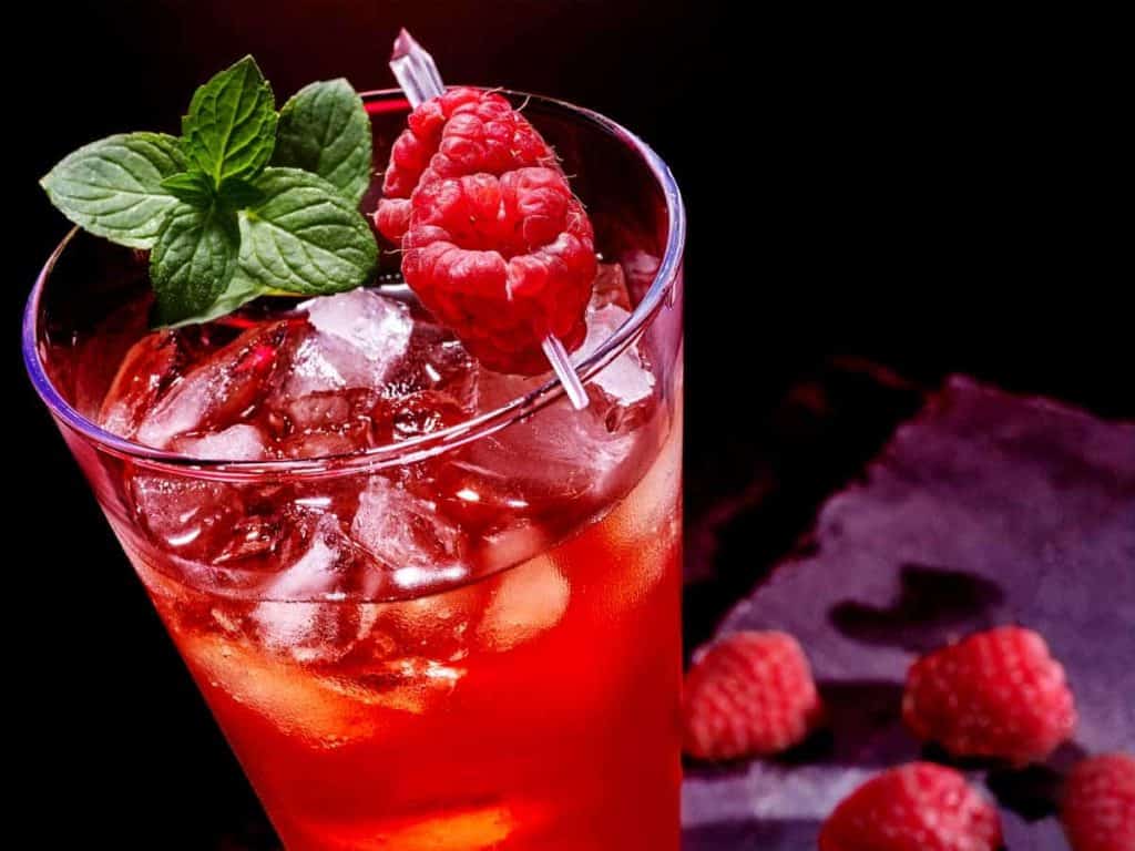 Ein Glas rotes Eisgetränk, garniert mit frischen Minzblättern und Himbeeren, mit zusätzlichen Himbeeren und einer dunklen Fläche im Hintergrund.