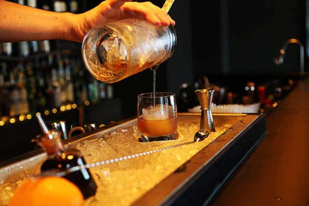 Eine Hand gießt einen braunen Cocktail aus einem Mixglas in ein Whiskyglas auf Eis, während auf der Theke Barutensilien und Zitrusfrüchte zu sehen sind.