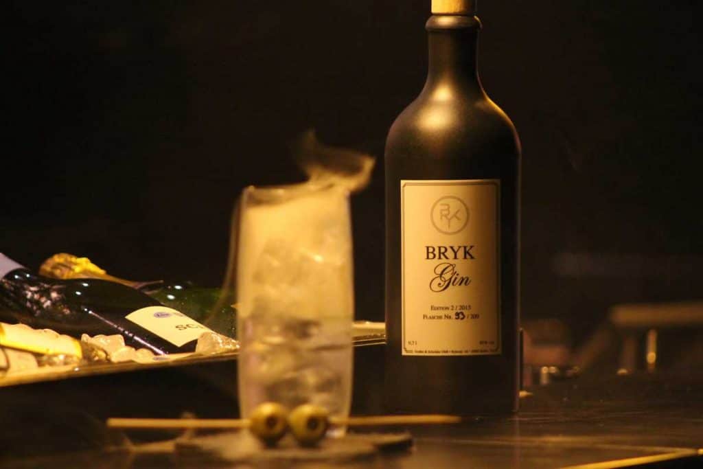 Auf einer schummrig beleuchteten Bar steht eine Flasche BRYK Gin neben einem mit Eis gefüllten Cocktailglas, das mit zwei Oliven garniert ist, während aus dem Getränk ein rauchiger Effekt aufsteigt.