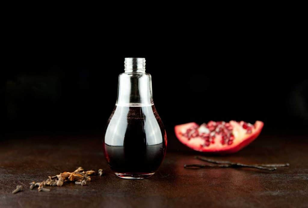Eine Glasflasche in Form einer Glühbirne, gefüllt mit einer dunklen Flüssigkeit, steht auf einer braunen Unterlage neben einem Granatapfelstück, zwei Vanilleschoten und Gewürzen – alles vor einem schwarzen Hintergrund.