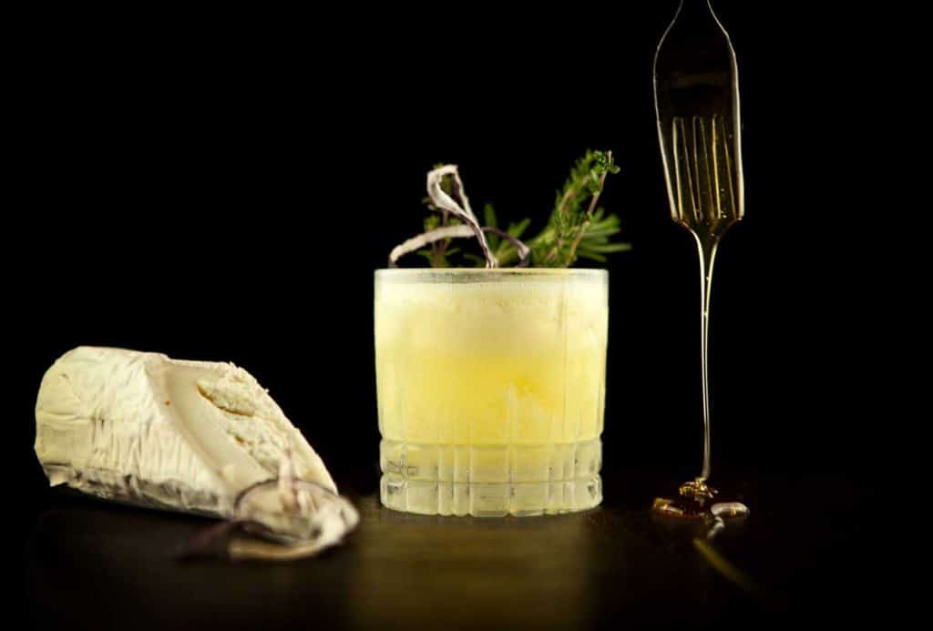 Ein Glas mit einem gelben Cocktail, garniert mit Kräutern, ein Stück Käse und eine Gabel, von der Honig tropft, sind auf einer dunklen Fläche vor schwarzem Hintergrund angeordnet.