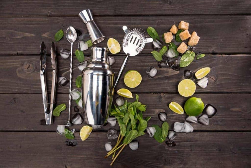 Auf einer dunklen Holzfläche liegen ein Cocktailshaker, ein Sieb, ein Jigger, ein Barlöffel, eine Zange, Minzblätter, Limettenschnitze, Zuckerwürfel und Eiswürfel.