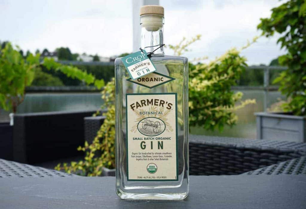 Auf einem Terrassentisch im Freien steht eine Flasche „Farmers Organic Gin“, im Hintergrund sind Grünflächen, ein See und Gartenstühle zu sehen.