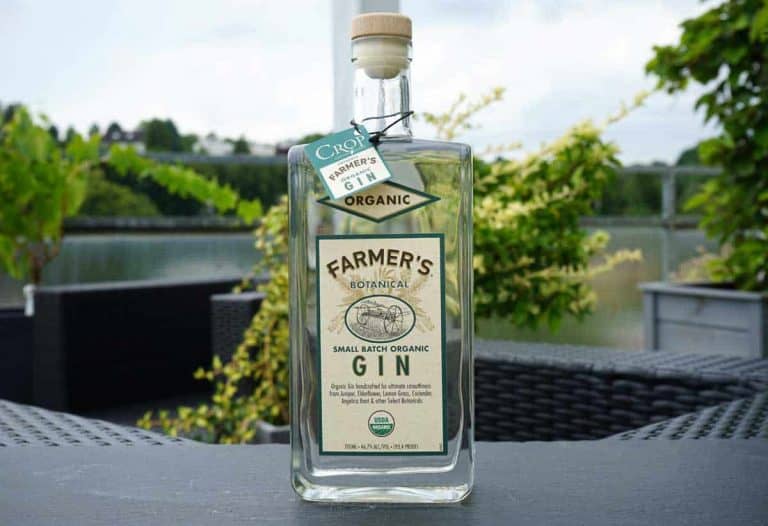 Auf einem Terrassentisch im Freien steht eine Flasche „Farmers Organic Gin“, im Hintergrund sind Grünflächen, ein See und Gartenstühle zu sehen.