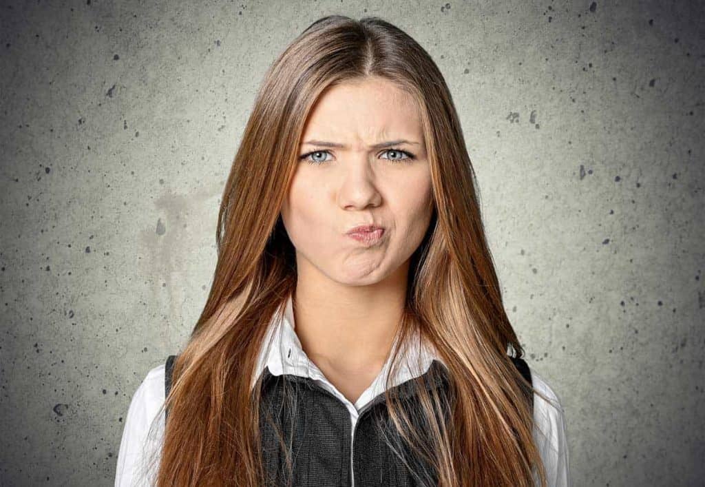 Eine Frau mit langen braunen Haaren macht einen verwirrten oder skeptischen Gesichtsausdruck, spitzt die Lippen und runzelt die Stirn, während sie vor einem grauen, strukturierten Hintergrund steht.