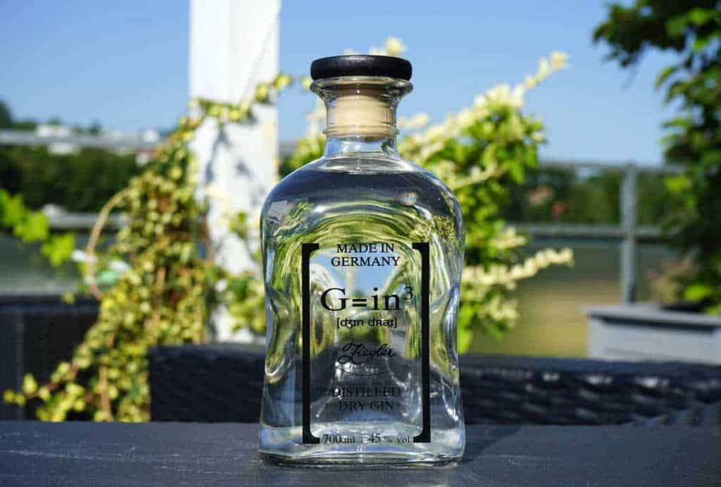Auf einem Tisch im Freien steht eine klare Glasflasche mit deutschem Gin und einem schwarzen Verschluss. Im Hintergrund sind unter blauem Himmel grüne Pflanzen und ein verschwommenes Balkongeländer zu sehen.