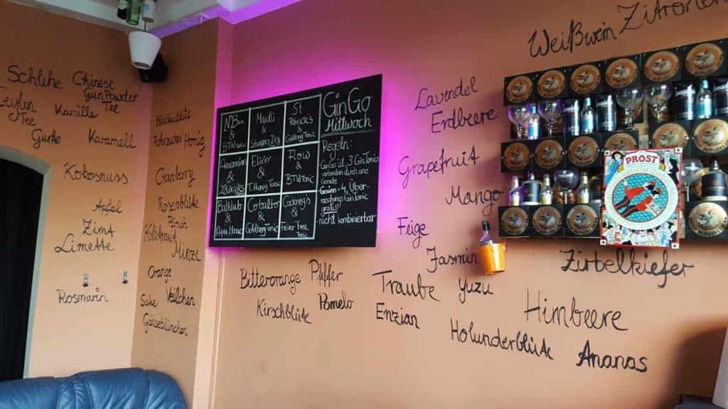 Eine Barwand mit handgeschriebenen Namen von Kräutern und Früchten auf Deutsch, ein Regal mit Gläsern, ein „Prost!“-Schild und eine Speisekarte auf einer Tafel, auf der verschiedene Gin-Tonic-Varianten aufgeführt sind.