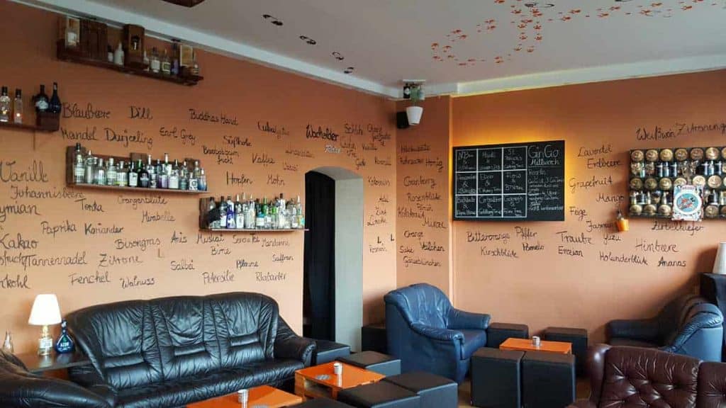Eine gemütliche Lounge mit schwarzen und blauen Ledersofas, Regalen voller Spirituosenflaschen, handgeschriebenen Namen an den orangefarbenen Wänden und einer Tafel mit der Getränkekarte.