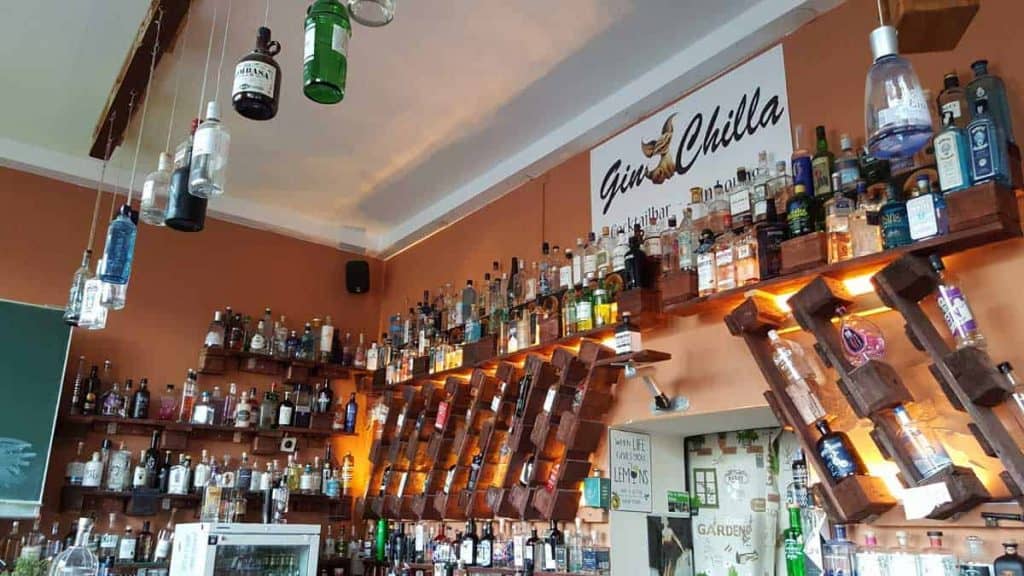 Eine Bar mit Regalen voller verschiedener Spirituosen, vor allem Gin-Flaschen, die ordentlich an der Wand aufgereiht sind. Einige Flaschen hängen kopfüber. Auf einem Schild steht „Gin Chilla“. Die Einrichtung besticht durch eine warme Beleuchtung und eine gemütliche Atmosphäre.