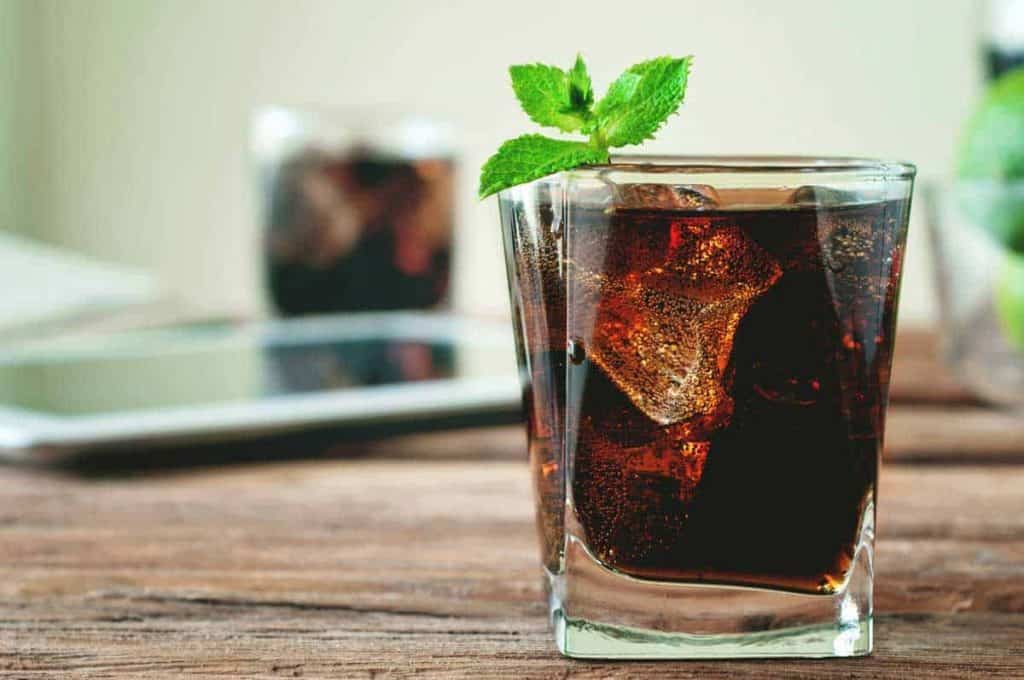 Auf einem Holztisch steht ein Glas Cola mit Eiswürfeln und einem Minzzweig darauf. Im Hintergrund sind ein weiteres Glas und verschwommene Gegenstände zu sehen.