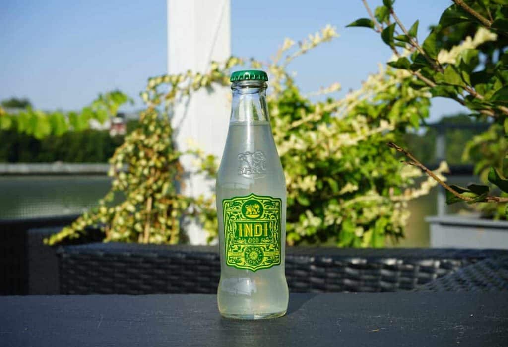 Auf einem schwarzen Tisch im Freien steht eine durchsichtige Glasflasche mit einem grün-gelben Etikett, auf dem „INDI“ steht; im Hintergrund sind Pflanzen, ein weißer Pfosten und ein blauer Himmel zu sehen.