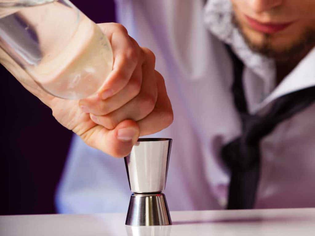 Nahaufnahme einer Person, die Flüssigkeit aus einer Flasche in einen Metall-Messbecher gießt; sie trägt ein weißes Hemd und eine schwarze Krawatte, wobei nur ein Teil ihres Gesichts zu sehen ist.