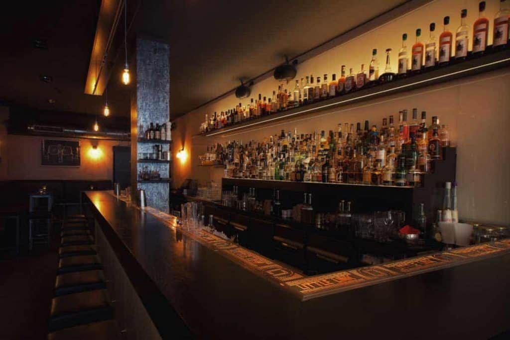 Eine schwach beleuchtete Bar mit Regalen voller Spirituosenflaschen hinter der Theke, schwarzen Barhockern entlang der Theke und warmen Hängeleuchten, die eine gemütliche, einladende Atmosphäre schaffen.