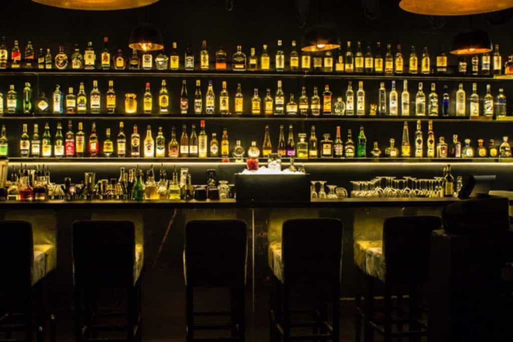 Eine schwach beleuchtete Bar mit Regalen voller verschiedener Spirituosenflaschen, Gläsern und vier Barhockern im Vordergrund. Warme Deckenbeleuchtung sorgt für eine gemütliche Atmosphäre.