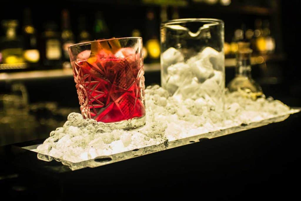 Auf einem Bett aus zerstoßenem Eis stehen an einer Bar ein Glas mit rotem Cocktail und ein Glas mit Eis, im Hintergrund sind verschiedene Flaschen verschwommen zu sehen.
