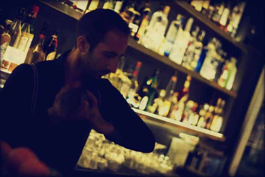 Ein Barkeeper steht hinter der Theke und schüttelt einen Cocktailshaker. Im Hintergrund sind Regale zu sehen, die mit Spirituosenflaschen und Gläsern gefüllt sind. Die Beleuchtung ist gedämpft und warm.