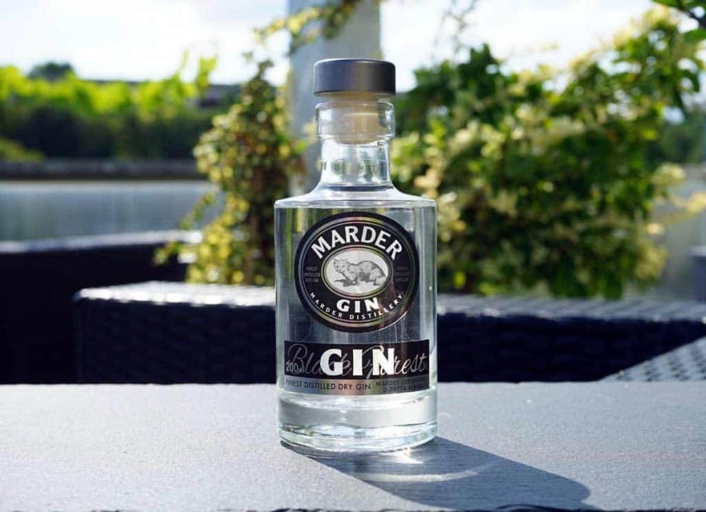 Auf einem dunklen Tisch im Freien steht eine klare Glasflasche mit Marder-Gin, im verschwommenen Hintergrund sind Grünpflanzen und Sonnenlicht zu sehen.