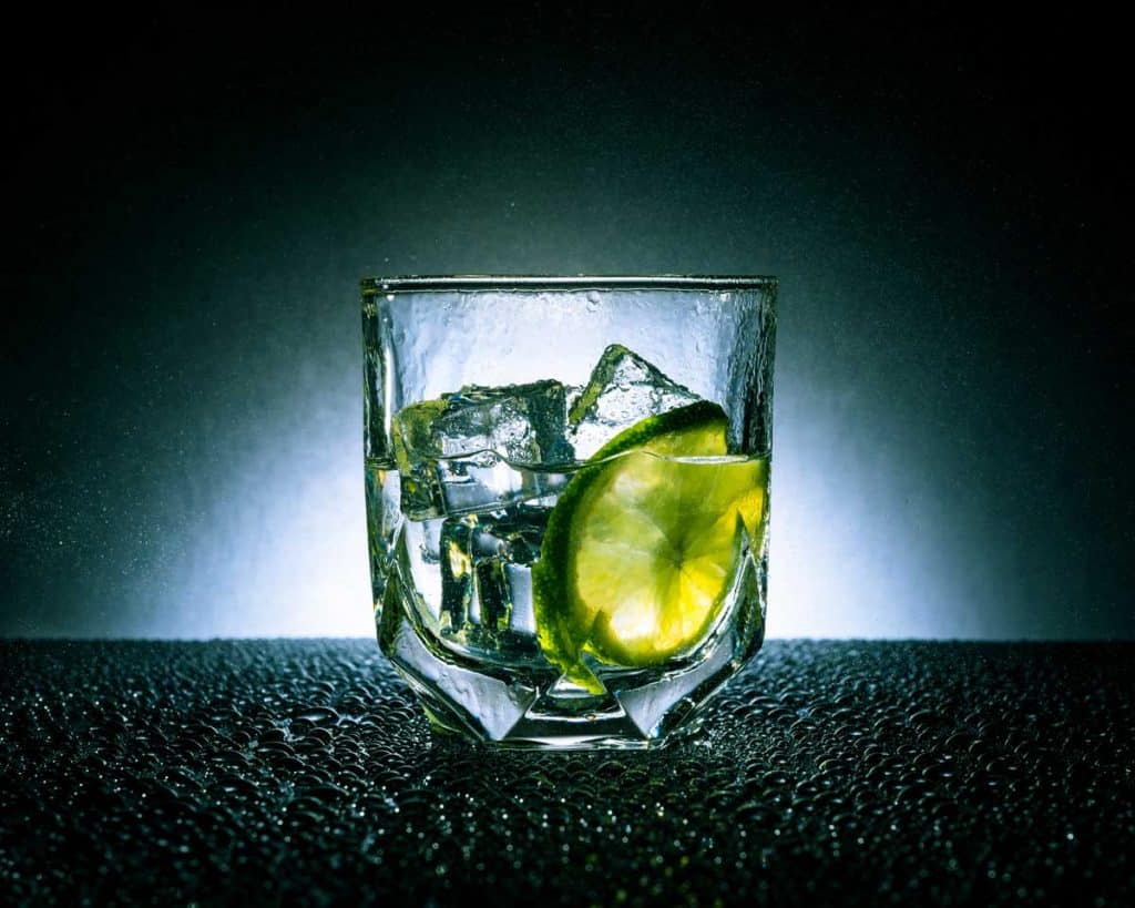 Ein mit Eiswürfeln und einer Limettenscheibe gefülltes Glas, das auf einer nassen Oberfläche vor einem dunklen Hintergrund und bei dramatischer Beleuchtung steht.