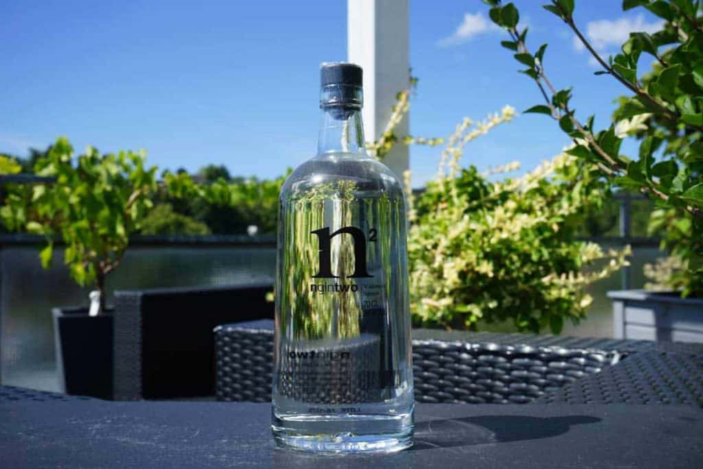 Eine durchsichtige Wasserflasche mit der Aufschrift „H2“ steht draußen auf einem dunklen Korbtisch, umgeben von grünen Pflanzen und vor dem Hintergrund eines blauen Himmels.