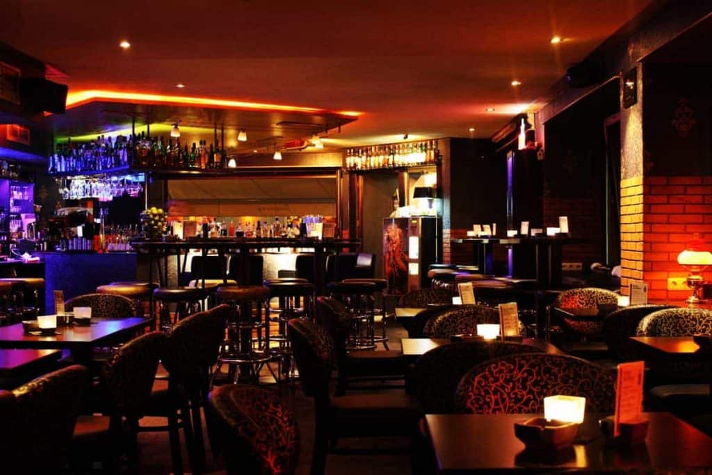 Eine schwach beleuchtete Bar mit gemusterten Stühlen, kleinen Tischen, leuchtenden Kerzen und einer gut sortierten Theke, an der die Flaschen in Reih und Glied stehen. Warme, stimmungsvolle Beleuchtung sorgt für eine gemütliche Atmosphäre.